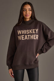 A woman stands against a plain background wearing the Panache Apparel Collection’s Dark Gray Whiskey Weather & Bourbon Weather Reversible Sweatshirt, featuring bold “WHISKEY WEATHER” lettering. She has long dark hair and is also dressed in dark pants.