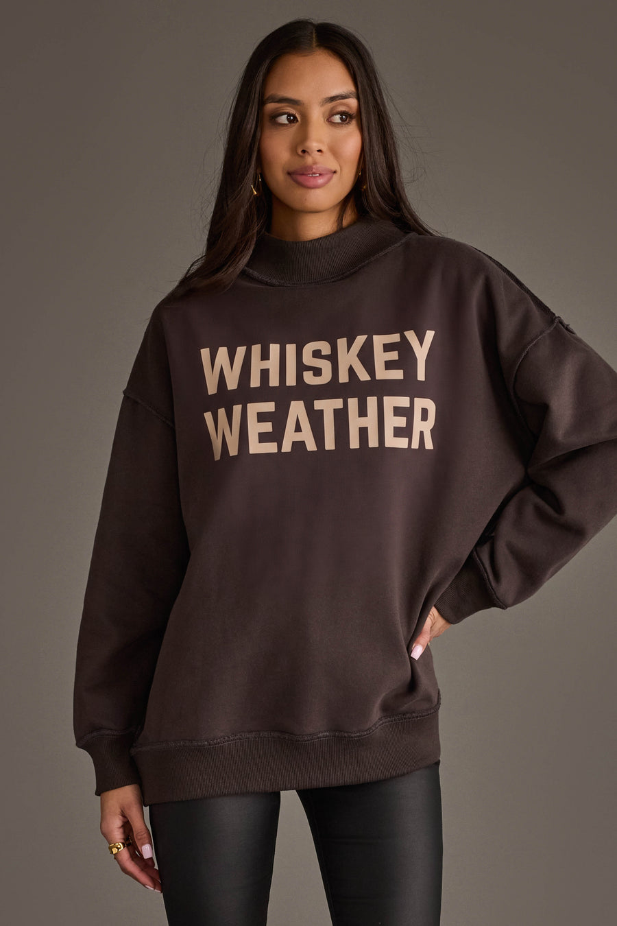 A woman stands against a plain background wearing the Panache Apparel Collection’s Dark Gray Whiskey Weather & Bourbon Weather Reversible Sweatshirt, featuring bold “WHISKEY WEATHER” lettering. She has long dark hair and is also dressed in dark pants.