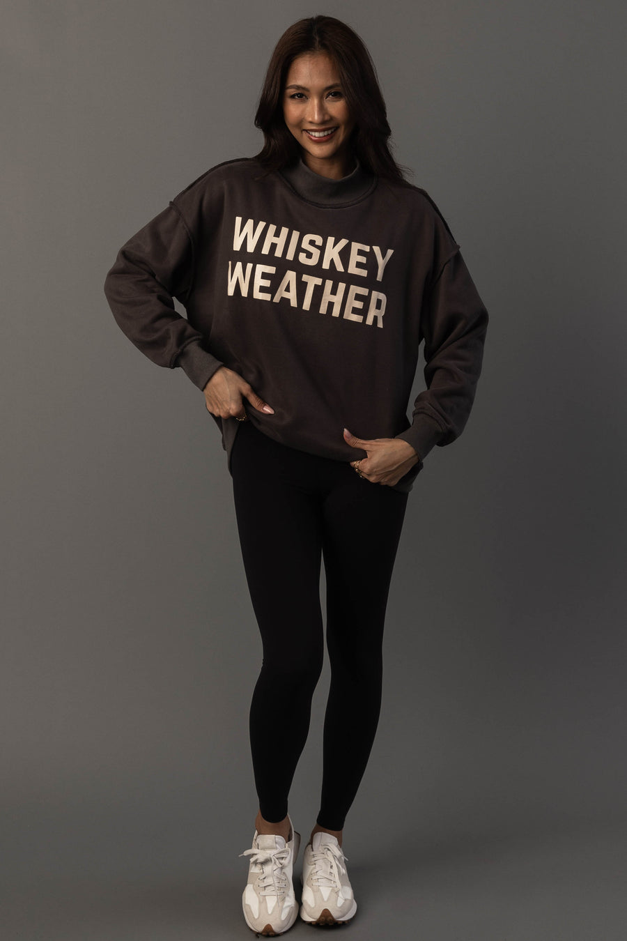 A smiling woman stands against a gray backdrop, wearing the Panache Apparel Collection’s Dark Gray Whiskey Weather & Bourbon Weather Reversible Sweatshirt, paired with black leggings and white sneakers.