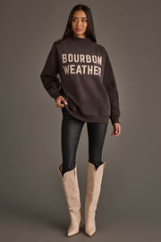 A woman stands against a gray background, wearing the Panache Apparel Collection’s Dark Gray Whiskey Weather & Bourbon Weather Reversible Sweatshirt along with black leggings and cream-colored cowboy boots. Her long dark hair cascades as she looks slightly to the side, highlighting her cozy Panache Bourbon Weather style.