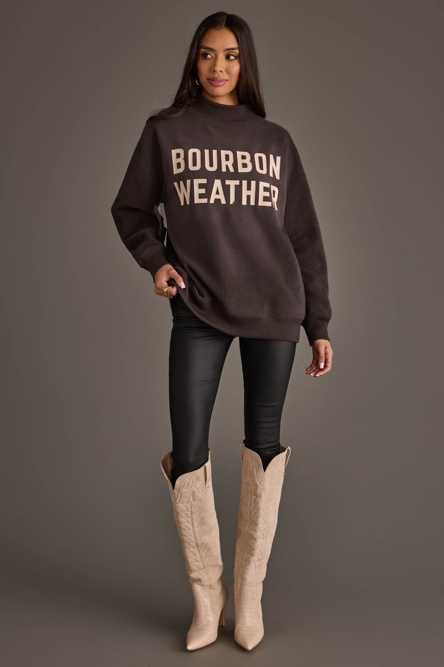 A woman stands against a gray background, wearing the Panache Apparel Collection’s Dark Gray Whiskey Weather & Bourbon Weather Reversible Sweatshirt along with black leggings and cream-colored cowboy boots. Her long dark hair cascades as she looks slightly to the side, highlighting her cozy Panache Bourbon Weather style.