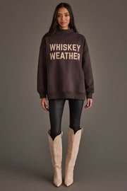 A woman stands against a plain background wearing the Dark Gray Whiskey Weather & Bourbon Weather Reversible Sweatshirt by Panache Apparel Collection, styled with bold lettering, black pants, and light-colored cowboy boots. Facing forward with a relaxed expression and both arms at her sides, she effortlessly highlights the cozy versatility of this reversible sweatshirt.