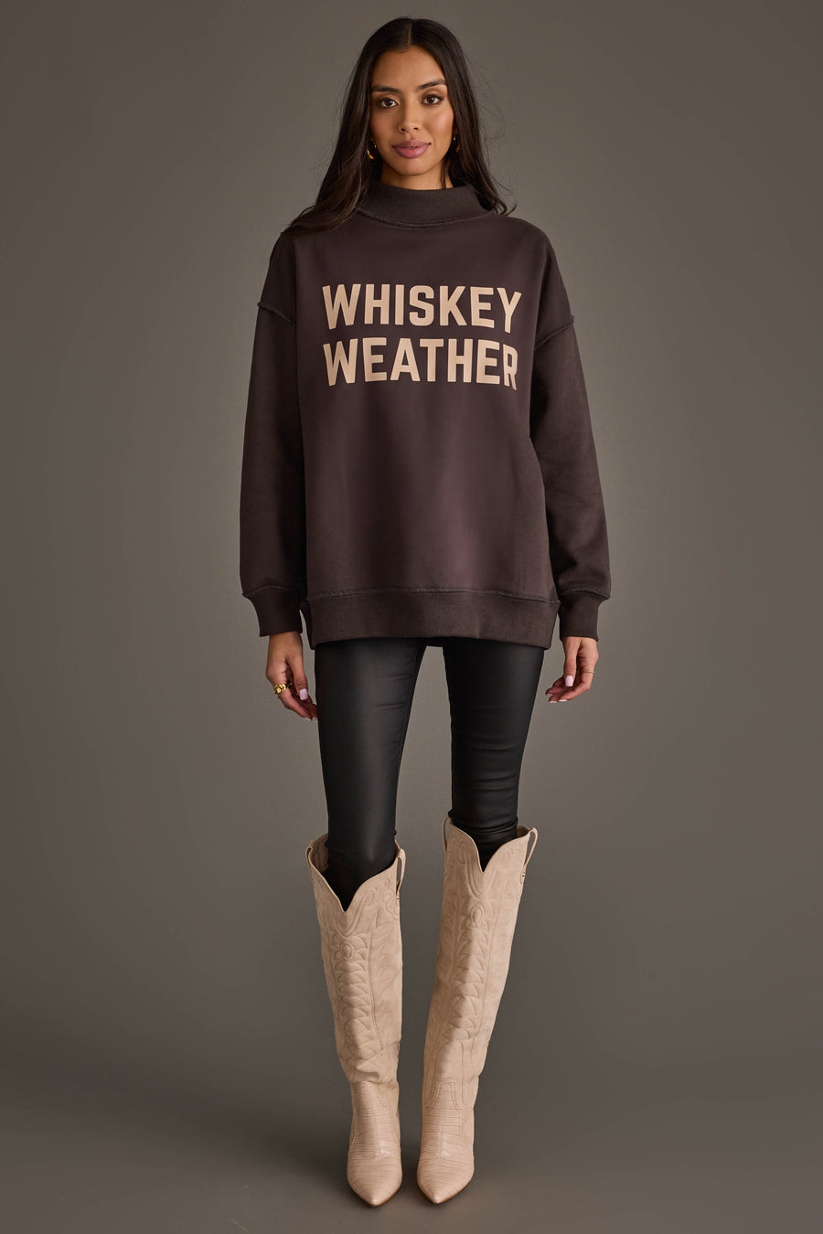 A woman stands against a plain background wearing the Dark Gray Whiskey Weather & Bourbon Weather Reversible Sweatshirt by Panache Apparel Collection, styled with bold lettering, black pants, and light-colored cowboy boots. Facing forward with a relaxed expression and both arms at her sides, she effortlessly highlights the cozy versatility of this reversible sweatshirt.