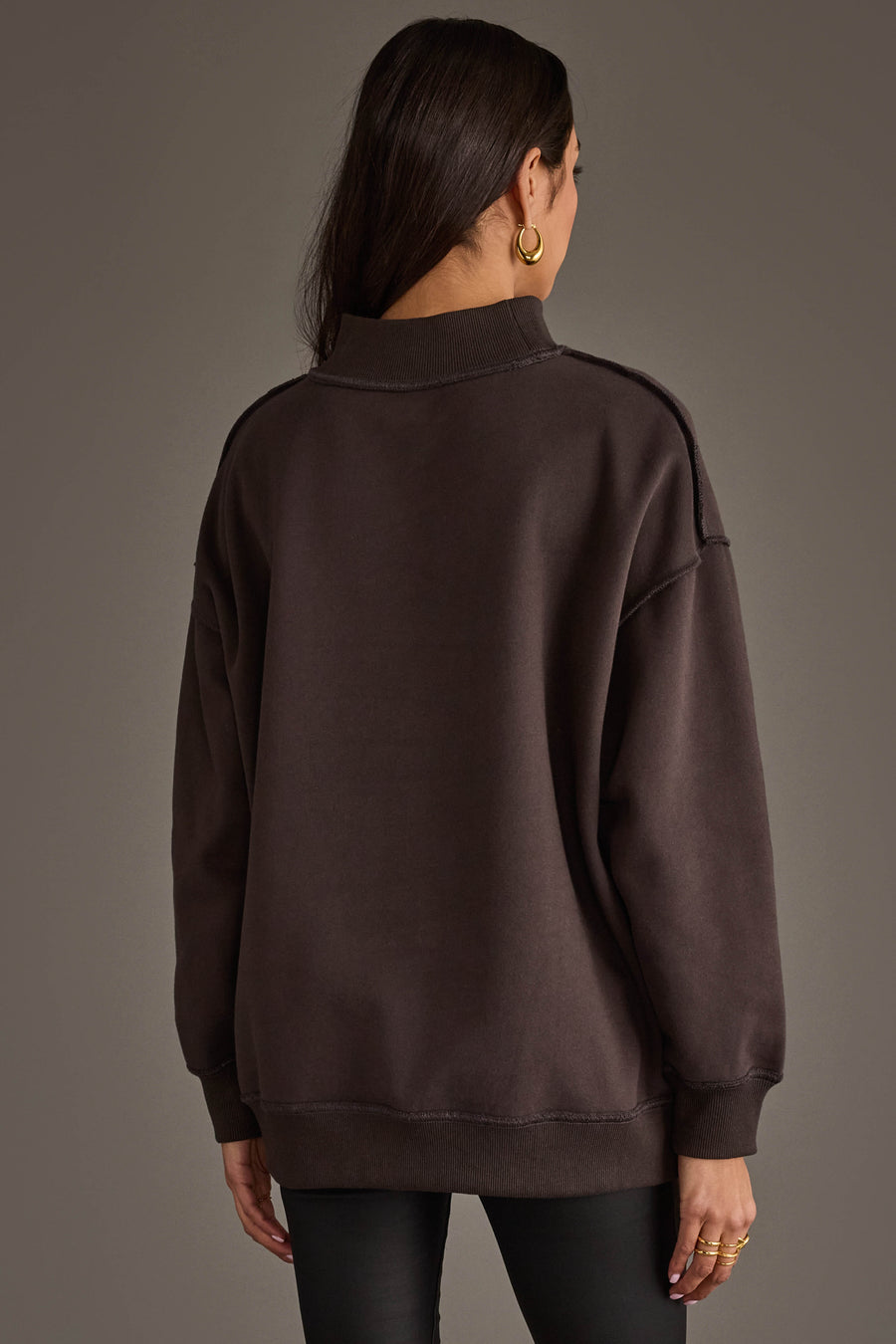 A person with long dark hair, wearing gold hoop earrings and the Panache Apparel Collection’s Dark Gray Day Drinking & Hungover Reversible Sweatshirt, stands facing away from the camera against a plain brown background.