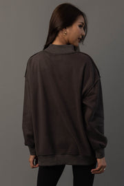 A woman with long brown hair stands facing away, wearing the Panache Apparel Collection Dark Gray Day Drinking & Hungover Reversible Sweatshirt and black pants, looking over her shoulder against a plain gray background.