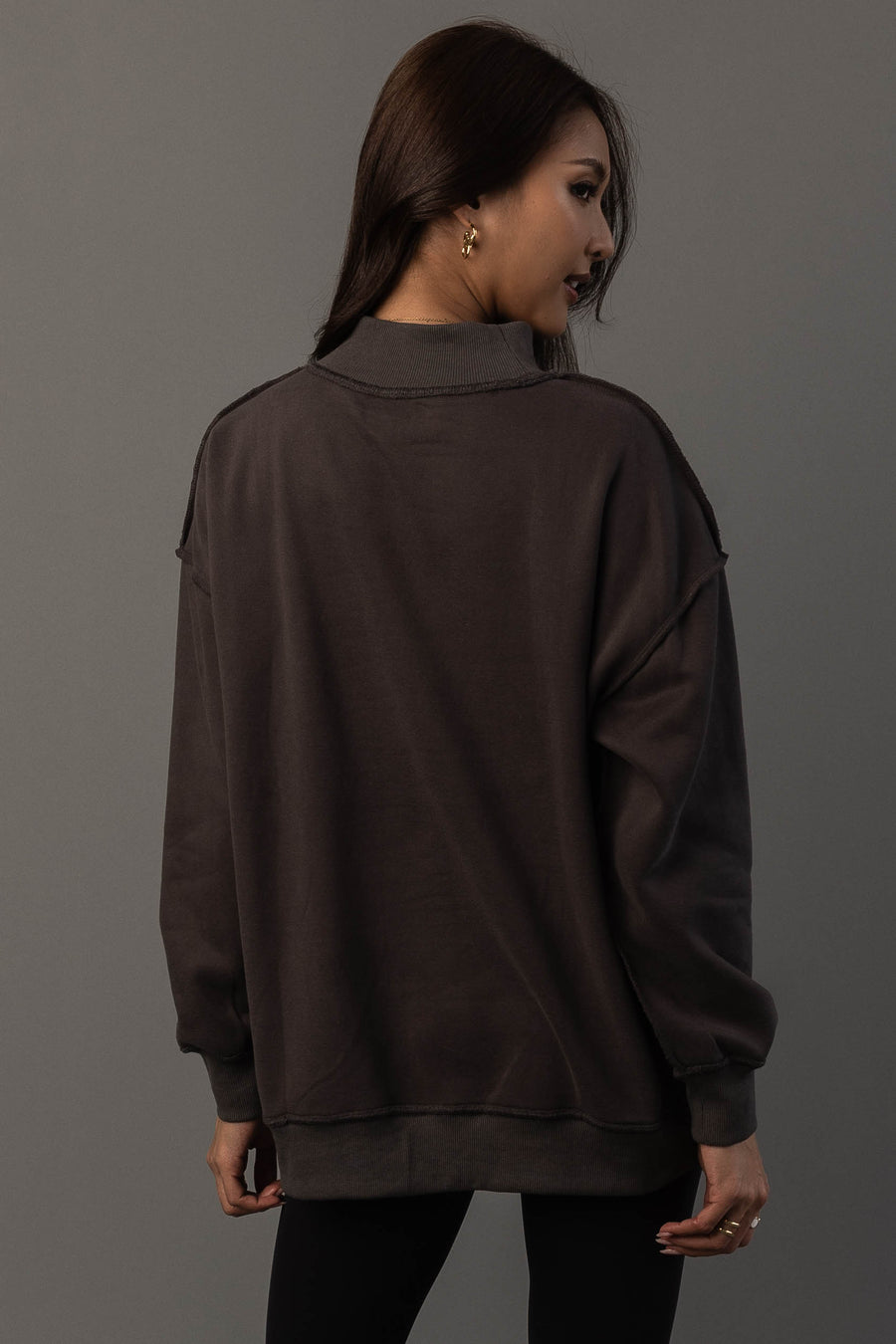 A woman with long brown hair stands facing away, wearing the Panache Apparel Collection Dark Gray Day Drinking & Hungover Reversible Sweatshirt and black pants, looking over her shoulder against a plain gray background.