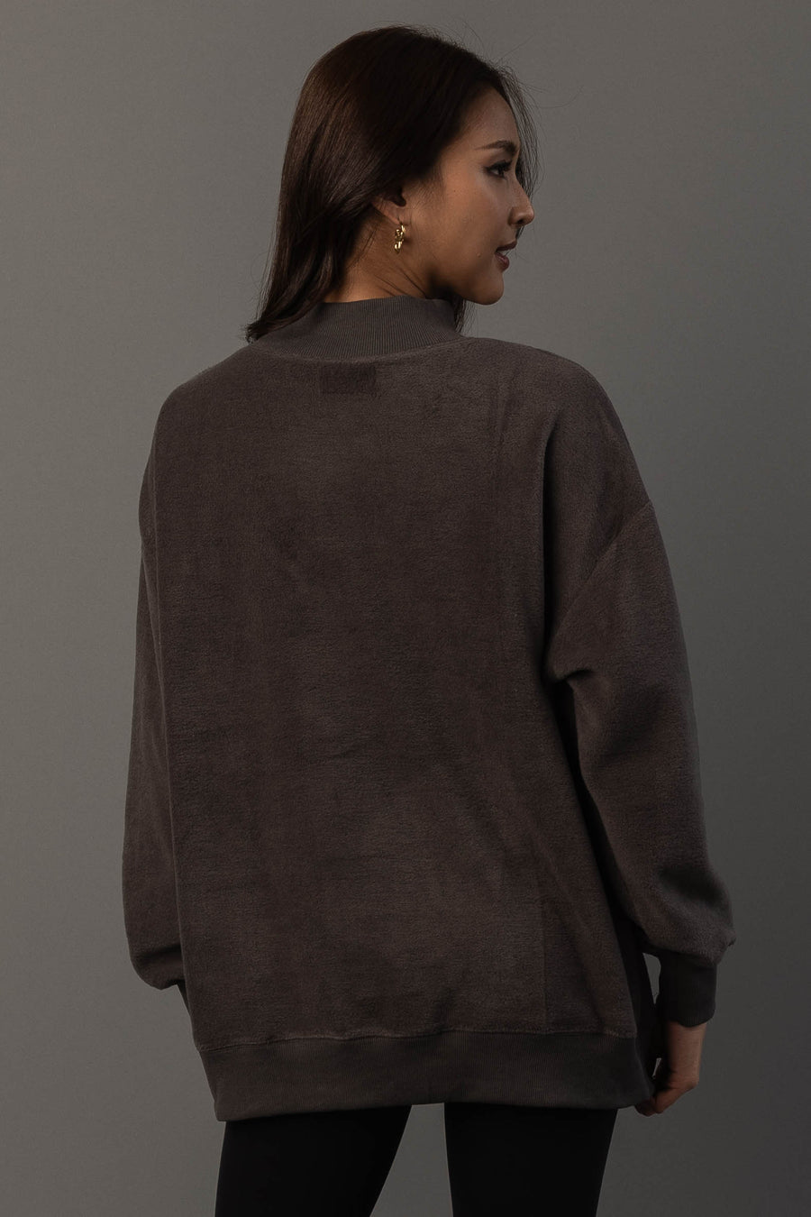 A woman with long brown hair, wearing gold earrings and the Panache Apparel Collection’s Dark Gray Day Drinking & Hungover Reversible Sweatshirt, stands facing away against a plain gray background.