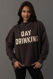 A woman with long brown hair wears the Panache Apparel Collection’s Dark Gray Day Drinking & Hungover Reversible Sweatshirt, which features large white lettering. She stands against a plain gray background, facing forward with a slight smile.