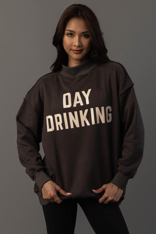 A woman with long brown hair wears the Panache Apparel Collection’s Dark Gray Day Drinking & Hungover Reversible Sweatshirt, which features large white lettering. She stands against a plain gray background, facing forward with a slight smile.