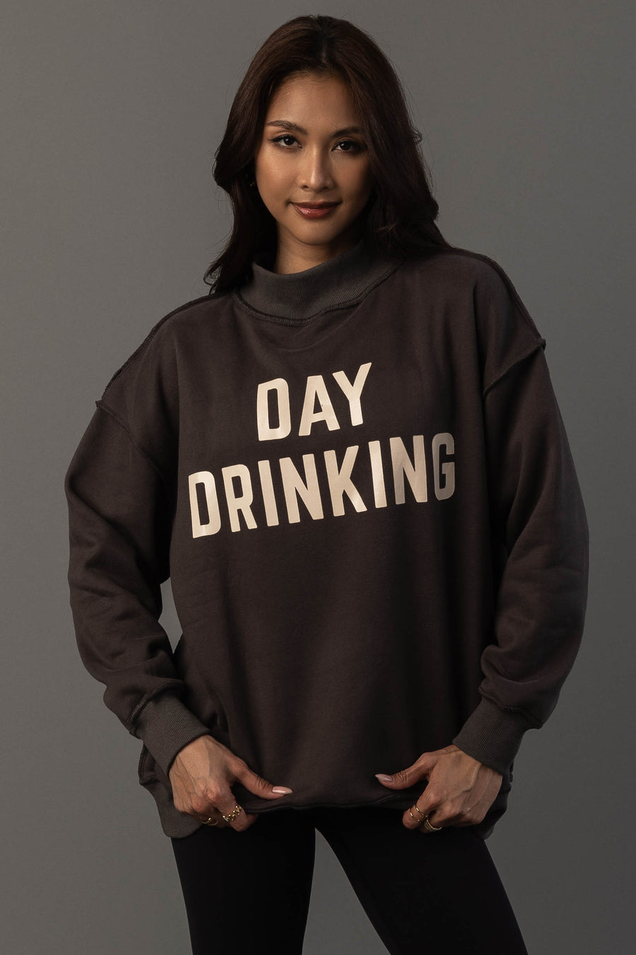 A woman with long brown hair wears the Panache Apparel Collection’s Dark Gray Day Drinking & Hungover Reversible Sweatshirt, which features large white lettering. She stands against a plain gray background, facing forward with a slight smile.