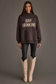 A woman stands against a gray background, wearing an oversized Panache Apparel Collection Dark Gray Day Drinking & Hungover Reversible Sweatshirt, paired with black leggings and tall beige cowboy boots.
