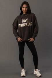 A woman stands against a gray backdrop wearing the Panache Apparel Collection’s Dark Gray Day Drinking & Hungover Reversible Sweatshirt, paired with black leggings and white sneakers. She faces forward, smiling slightly, with her hands touching her sweatshirt.