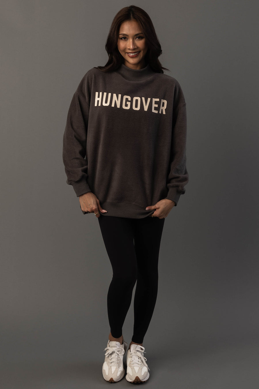 A woman stands against a gray background, smiling and wearing the Panache Apparel Collection Dark Gray Day Drinking & Hungover Reversible Sweatshirt, paired with black leggings and white sneakers.