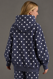 A person with blonde hair is seen from behind, standing against a plain gray background and wearing the Panache Apparel Collection Navy & White Polka Dot Hooded Sweatshirt, styled with matching pants featuring large white polka dots.