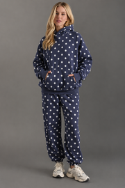 A woman models the Panache Apparel Collection’s Navy & White Polka Dot Hooded Sweatshirt, paired with matching sweatpants and white sneakers, with her hands in her pockets against a plain gray background.