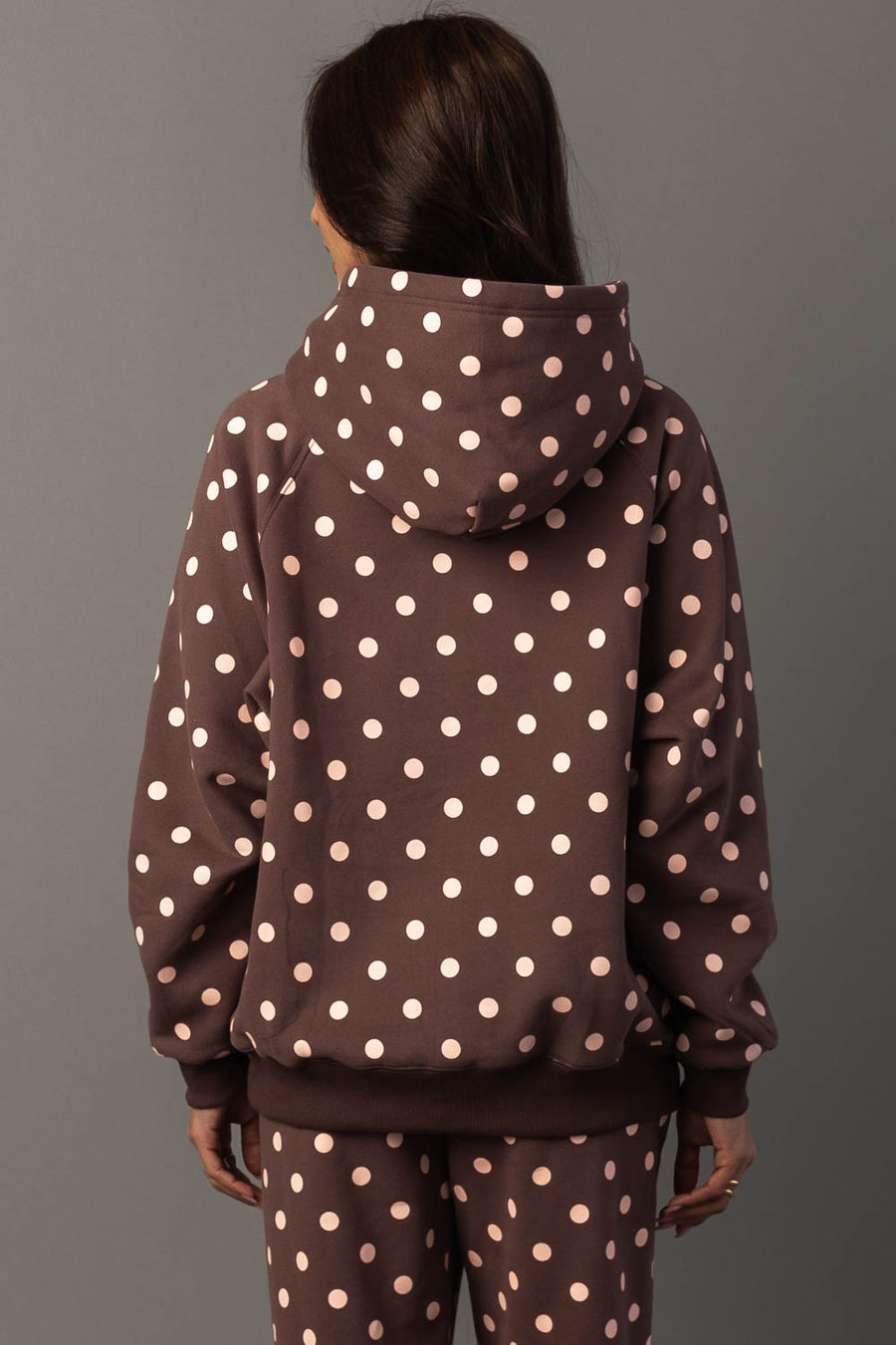 A person with long brown hair, seen from behind, is wearing the Panache Apparel Collection Brown & Light Pink Polka Dot Hooded Sweatshirt, standing against a plain gray background.