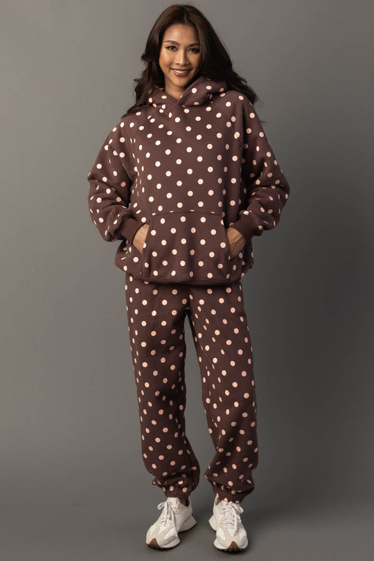 A woman smiles while wearing the Panache Apparel Collection’s Brown & Light Pink Polka Dot Hooded Sweatshirt, paired with matching sweatpants and white sneakers, standing against a plain gray background.
