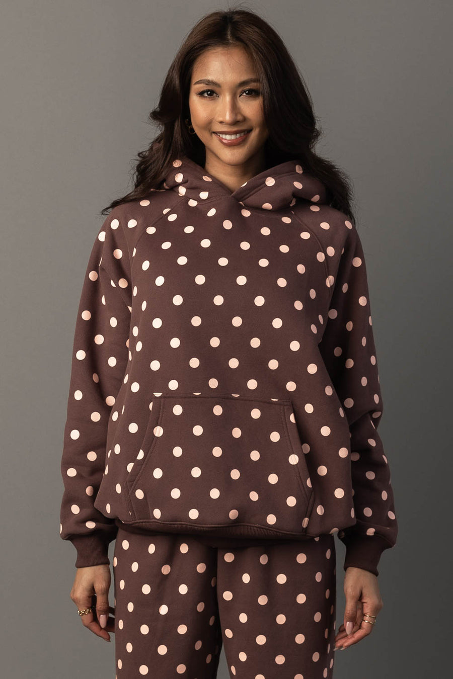 A woman stands smiling, wearing the Panache Apparel Collection Brown & Light Pink Polka Dot Hooded Sweatshirt, paired with matching pants, and poses against a plain gray background.