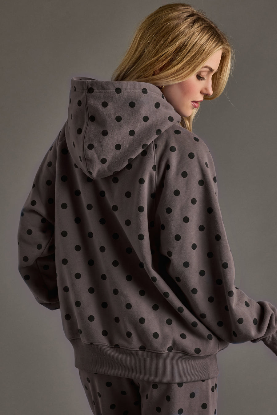A woman with blonde hair, seen from behind, wears the Panache Apparel Collection’s Dark Gray & Black Polka Dot Hooded Sweatshirt, paired with matching pants featuring black polka dots. The background is plain and neutral.