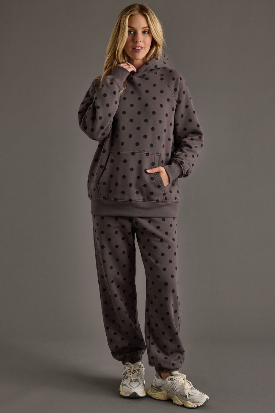 A person models the Panache Apparel Collection’s Dark Gray & Black Polka Dot Hooded Sweatshirt, paired with matching sweatpants, standing against a plain gray background with one hand near their chin and the other in their pocket.