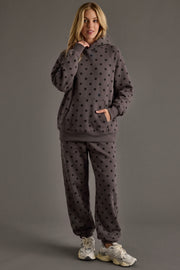 A person stands against a gray background, wearing Dark Gray & Black Polka Dot Sweatpants from Panache Apparel Collection along with a matching hooded sweatshirt and white sneakers. One hand is in the hoodie pocket, while the other lightly touches their chin.