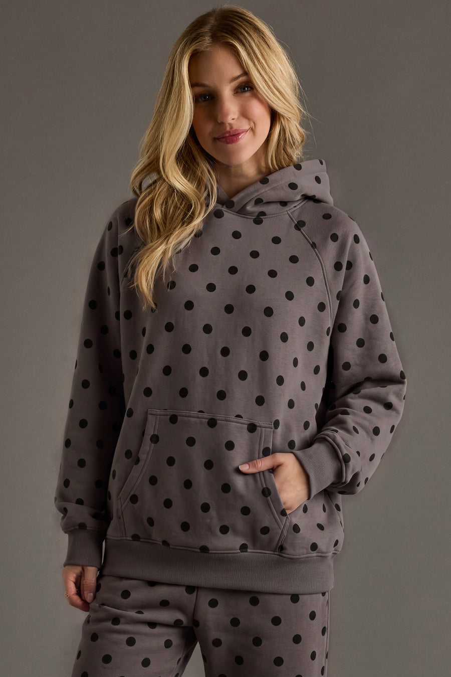 A woman with long blonde hair wears the Panache Apparel Collection Dark Gray & Black Polka Dot Hooded Sweatshirt and matching pants, standing against a plain gray background and smiling gently at the camera.
