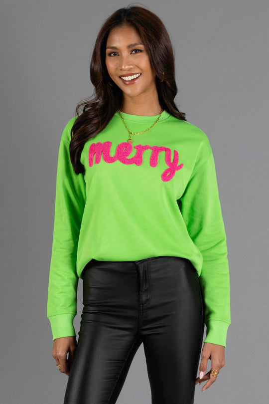 A woman wears a bright green Bright Green Merry Sweatshirt featuring the word merry in large, pink fuzzy script across the chest.