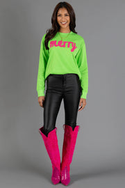 A smiling woman wears a Bright Green Merry Sweatshirt with pink lettering, paired with black pants and knee-high pink western boots.