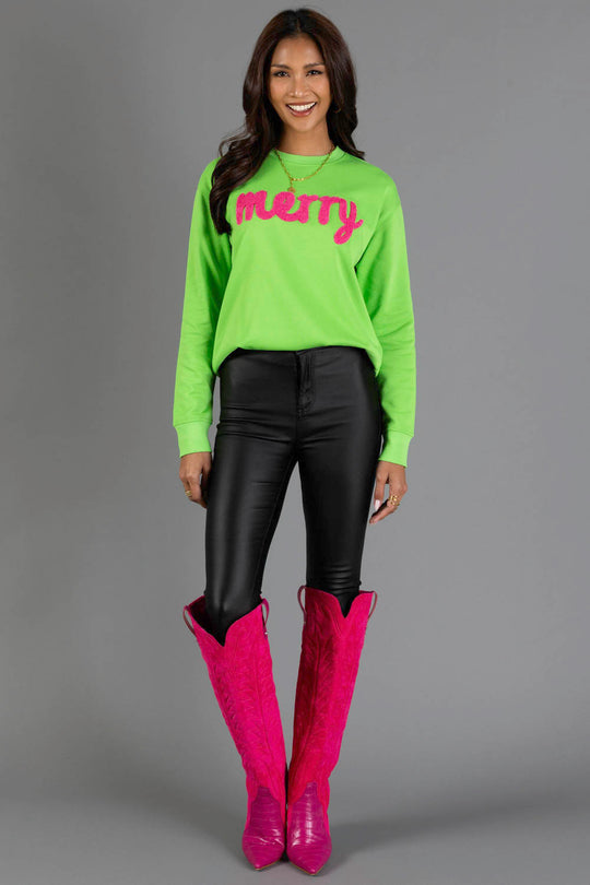 A smiling woman wears a Bright Green Merry Sweatshirt with pink lettering, paired with black pants and knee-high pink western boots.
