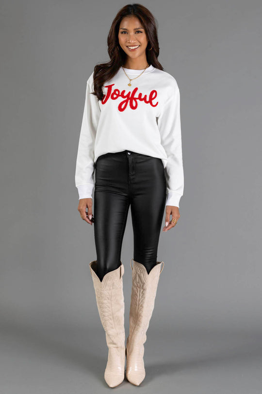 A woman wears a white Joyful Sweatshirt by Panache Apparel Collection with a crew neckline, paired with black pants and tall beige boots.