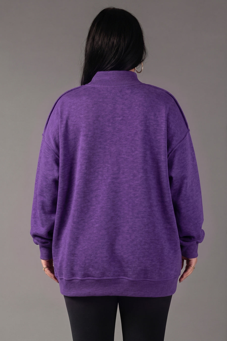 A person with long dark hair, wearing the Panache Apparel Collection Purple Game Day & Sunday Funday Reversible Sweatshirt and black pants, stands facing away from the camera against a plain gray background.