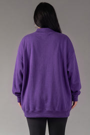 A person with long dark hair, wearing the Panache Apparel Collection’s Purple Game Day & Sunday Funday Reversible Sweatshirt and black pants, stands facing away from the camera against a plain gray background.