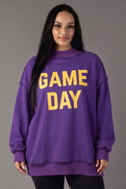 A woman with long dark hair, wearing the Panache Apparel Collection’s Purple Game Day & Sunday Funday Reversible Sweatshirt featuring bold yellow lettering, stands against a plain gray background and smiles at the camera.