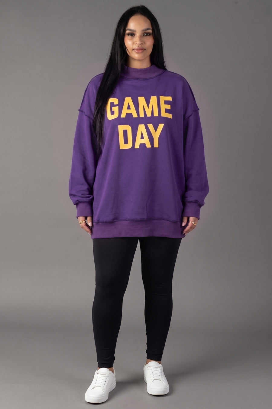 A woman stands against a gray background wearing the Panache Apparel Collection’s Purple Game Day & Sunday Funday Reversible Sweatshirt in bold yellow letters, paired with black leggings and white sneakers—creating that quintessential Sunday Funday vibe.