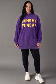 A woman stands facing forward, dressed in the Panache Apparel Collection Purple Game Day & Sunday Funday Reversible Sweatshirt with yellow lettering, styled with black leggings and white sneakers, against a plain gray background.