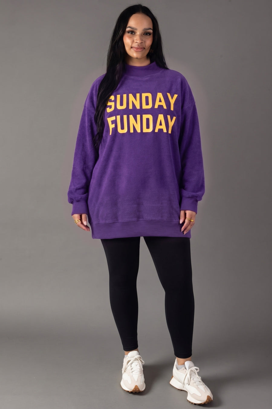 A woman stands facing forward, dressed in the Panache Apparel Collection Purple Game Day & Sunday Funday Reversible Sweatshirt with yellow lettering, styled with black leggings and white sneakers, against a plain gray background.