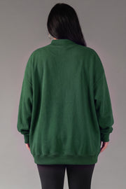 A person with long dark hair, wearing the Panache Apparel Collection’s Green Game Day & Sunday Funday Reversible Sweatshirt and black pants, is standing facing away from the camera against a plain gray background.