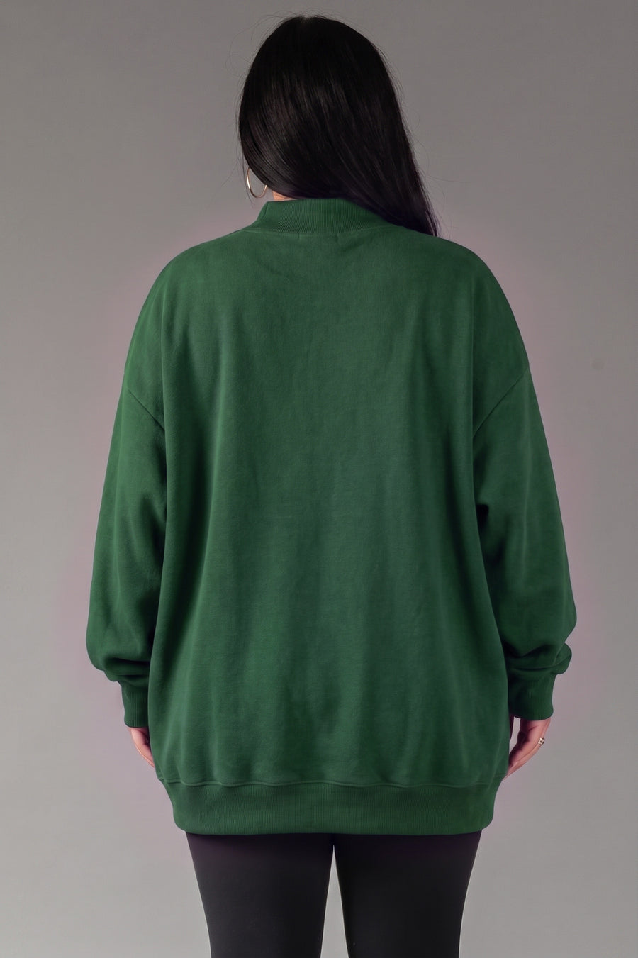 A person with long dark hair, wearing the Panache Apparel Collection’s Green Game Day & Sunday Funday Reversible Sweatshirt and black pants, is standing facing away from the camera against a plain gray background.