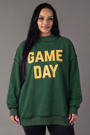 A woman with long dark hair stands and smiles against a plain gray background, wearing the Panache Apparel Collection’s Green Game Day & Sunday Funday Reversible Sweatshirt featuring bold yellow “GAME DAY” lettering.