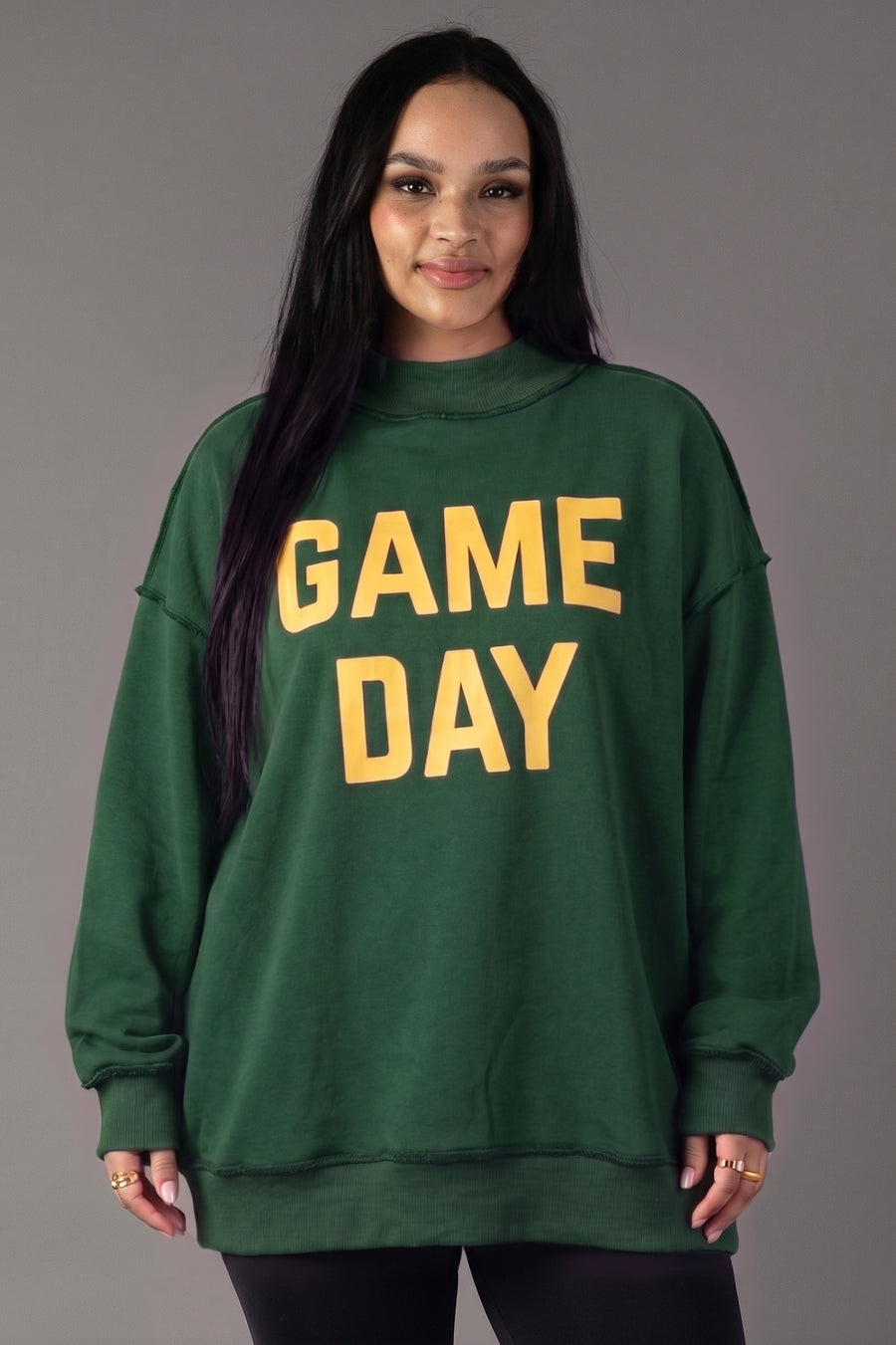 A woman with long dark hair stands and smiles against a plain gray background, wearing the Panache Apparel Collection’s Green Game Day & Sunday Funday Reversible Sweatshirt featuring bold yellow “GAME DAY” lettering.
