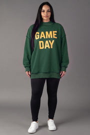 A person stands facing forward, wearing the Panache Apparel Collection Green Game Day & Sunday Funday Reversible Sweatshirt with yellow lettering, paired with black leggings and white sneakers, against a plain gray background.