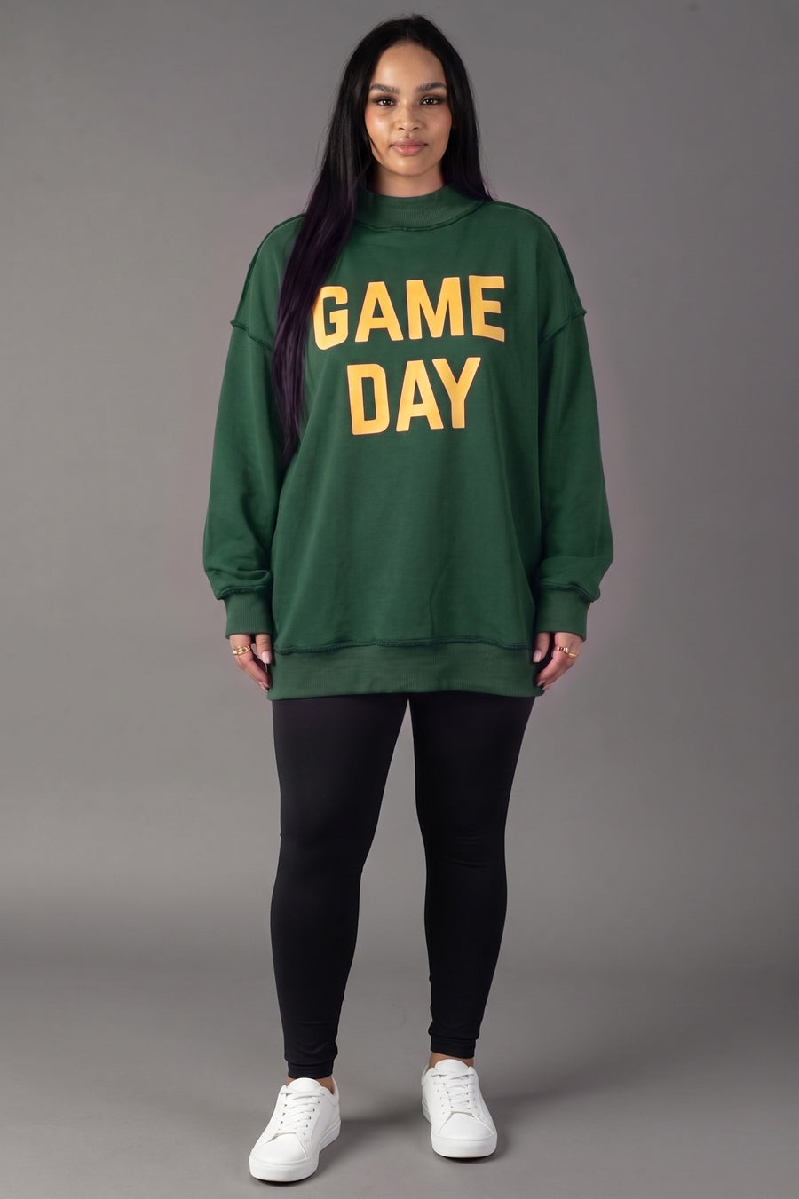 A person stands facing forward, wearing the Panache Apparel Collection Green Game Day & Sunday Funday Reversible Sweatshirt with yellow lettering, paired with black leggings and white sneakers, against a plain gray background.