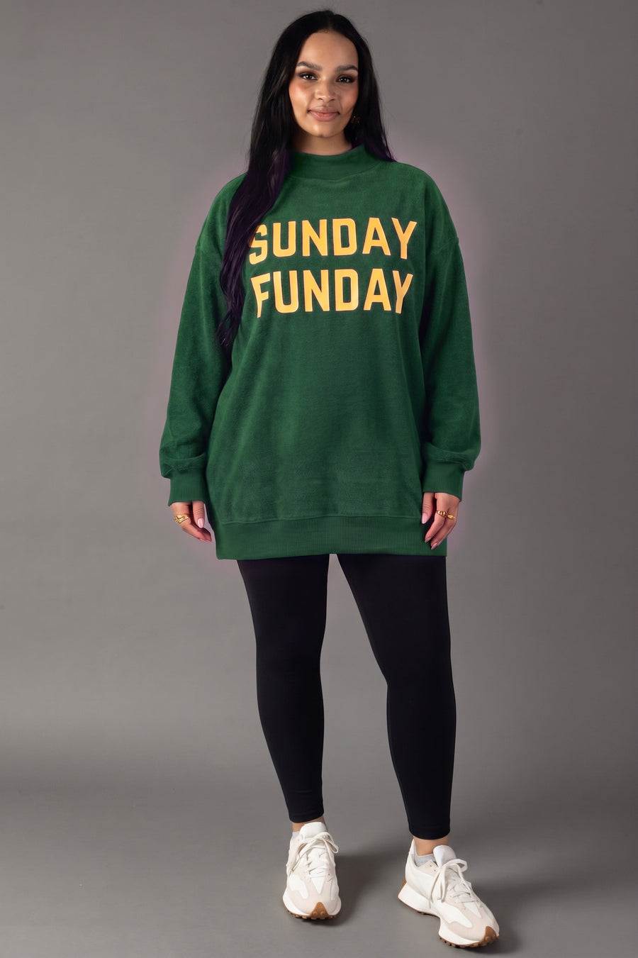 A person stands against a gray background wearing the Green Game Day & Sunday Funday Reversible Sweatshirt by Panache Apparel Collection, paired with black leggings and white sneakers.