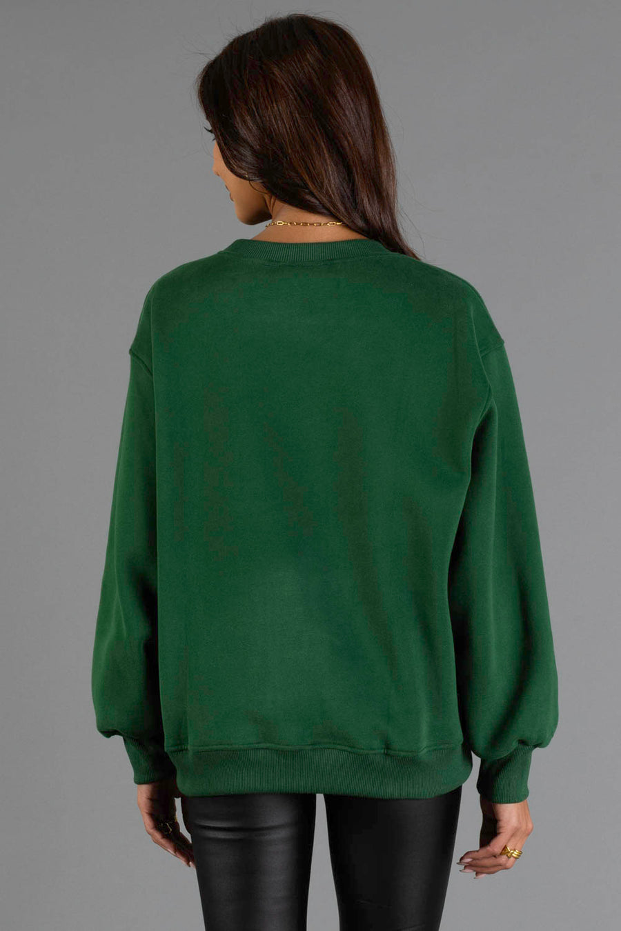 A woman with long brown hair, wearing the Panache Apparel Collection Green & Cream On Mountain Time Sweatshirt and black leather pants, is standing with her back to the camera against a plain gray background.
