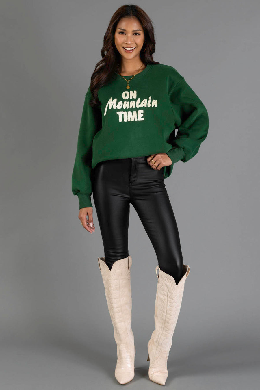 A smiling woman stands against a gray background wearing the Panache Apparel Collection Green & Cream On Mountain Time Sweatshirt, paired with black leather pants and tall white cowboy boots.