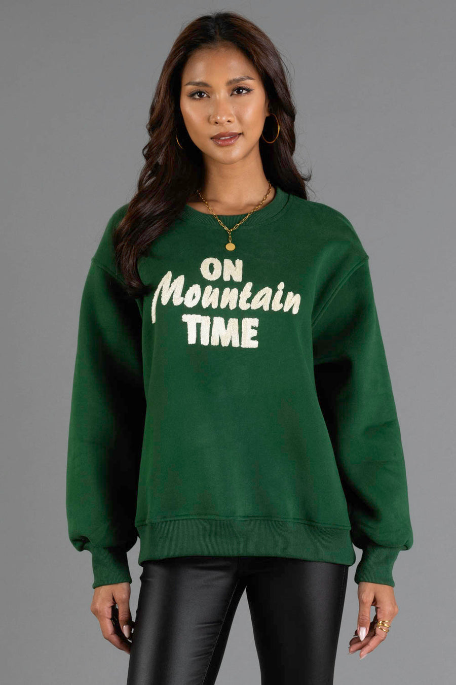 A woman wearing the Panache Apparel Collection Green & Cream On Mountain Time Sweatshirt, paired with black pants and gold jewelry, stands against a gray background.