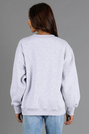 A person with long brown hair, wearing the Panache Apparel Collection’s “Heather Gray & Brown On Mountain Time” sweatshirt and blue jeans, stands facing away from the camera against a plain gray background.