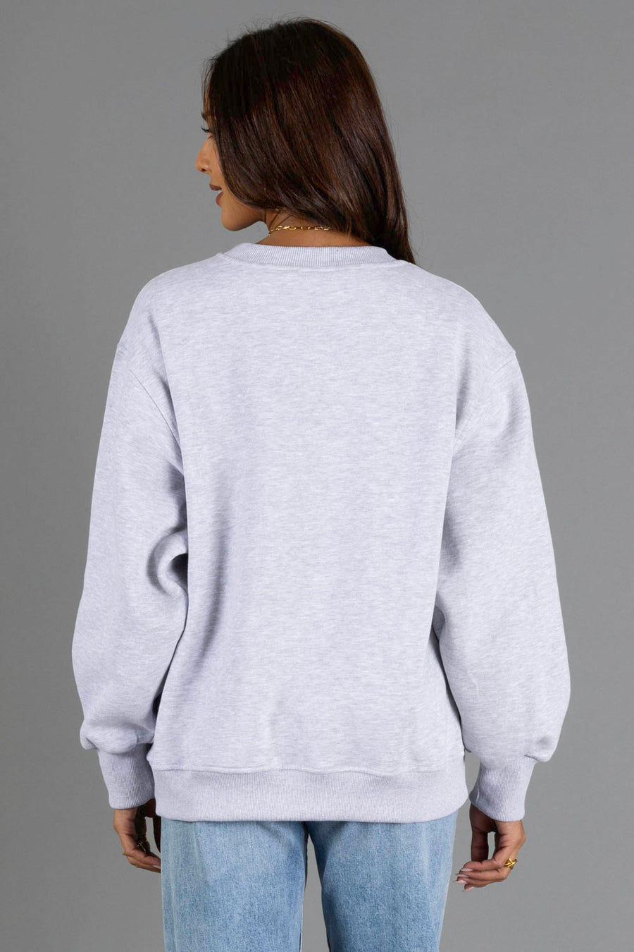 A person with long brown hair, wearing the Panache Apparel Collection’s “Heather Gray & Brown On Mountain Time” sweatshirt and blue jeans, stands facing away from the camera against a plain gray background.