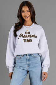 A woman with long brown hair smiles, wearing the Panache Apparel Collection Heather Gray & Brown On Mountain Time Sweatshirt and blue jeans, standing against a plain gray background.