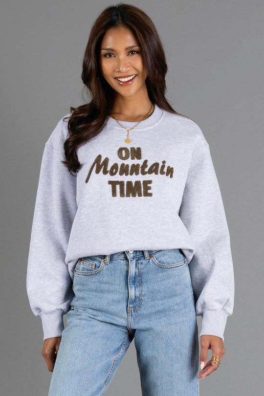 A woman with long brown hair smiles, wearing the Panache Apparel Collection Heather Gray & Brown On Mountain Time Sweatshirt and blue jeans, standing against a plain gray background.